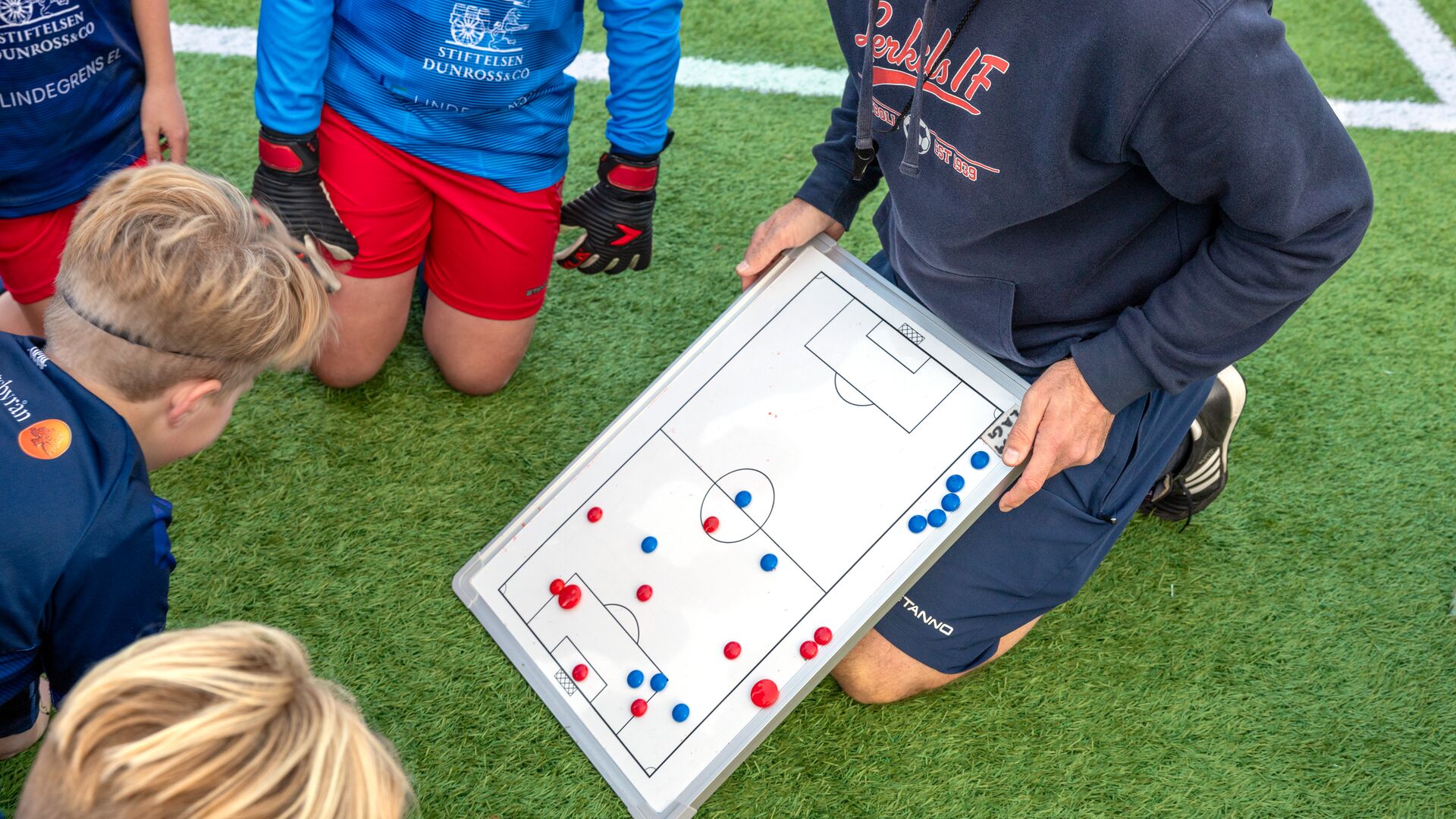 Lerkils tränare står på knä på fotbollsplanen och visar taktiktavla för laget