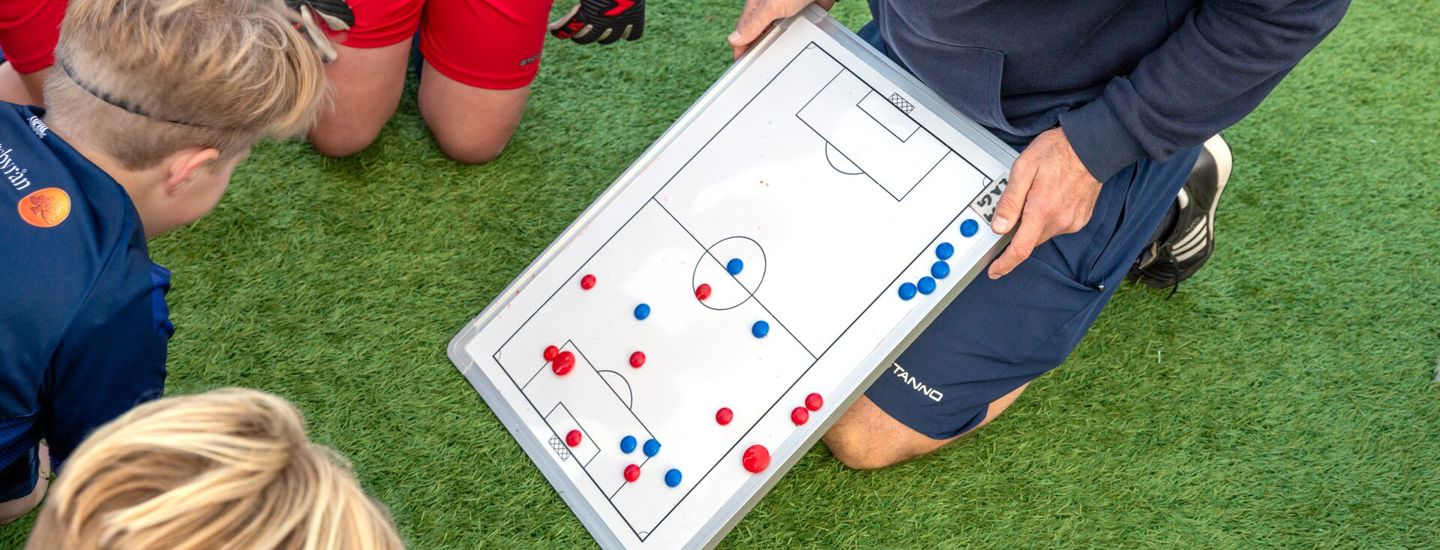Lerkils tränare står på knä på fotbollsplanen och visar taktiktavla för laget