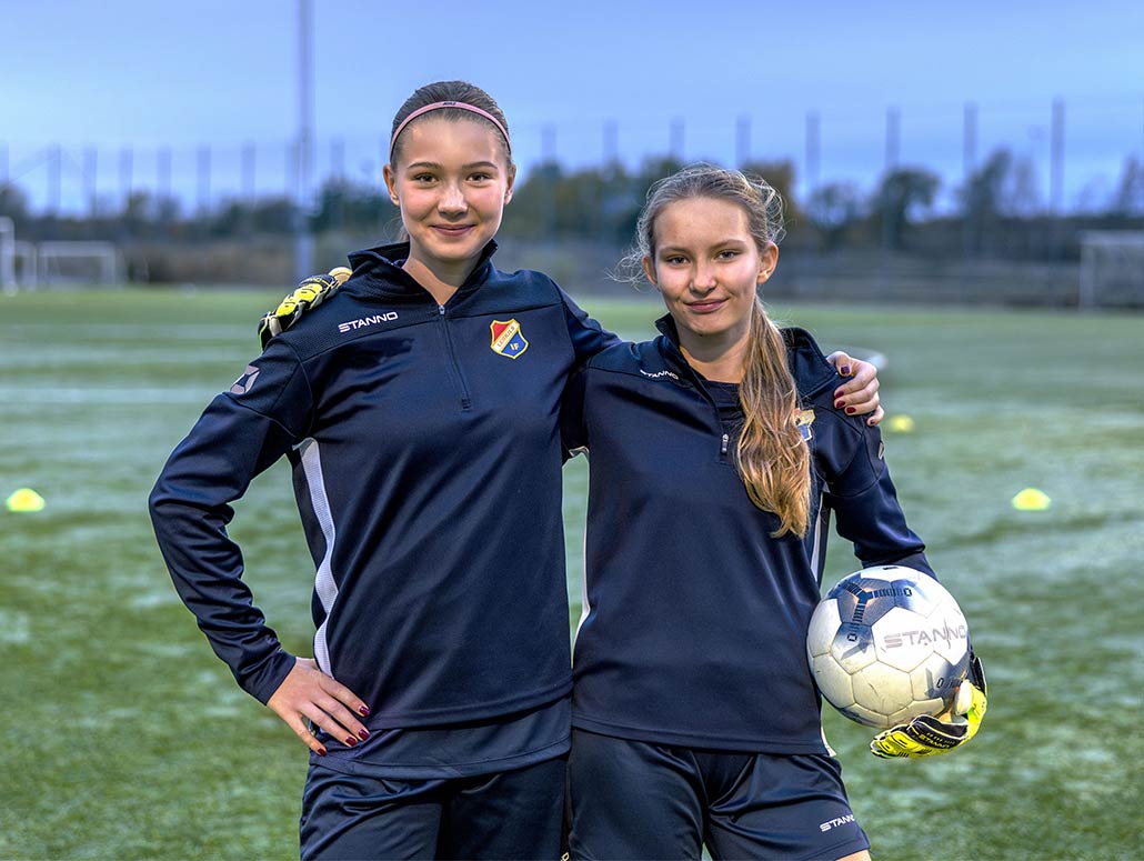 Två fotbollstjejer som kramar om varann och håller i en fotboll