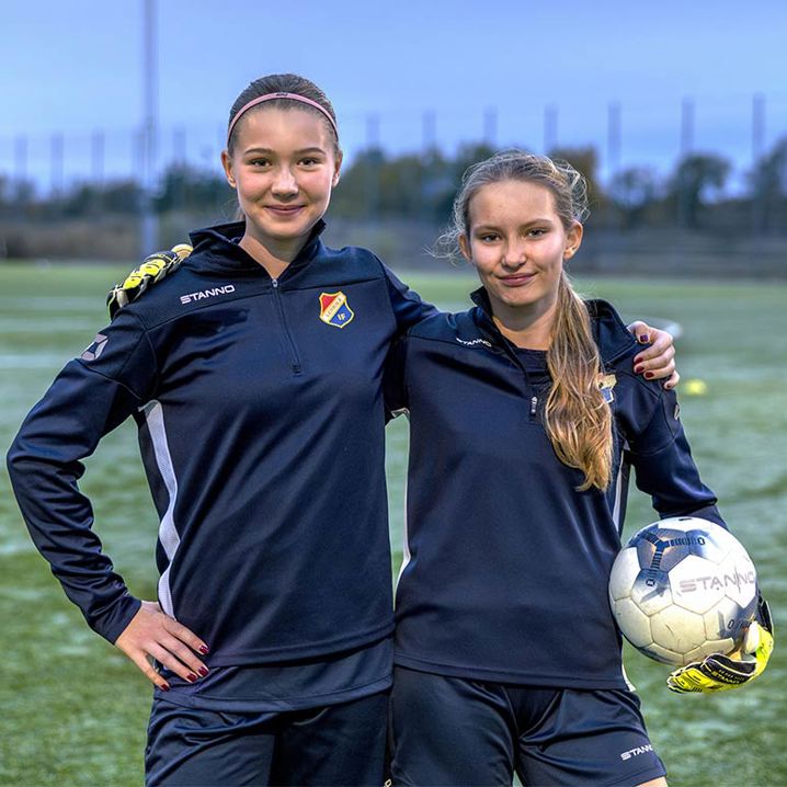 Två fotbollstjejer som kramar om varann och håller i en fotboll