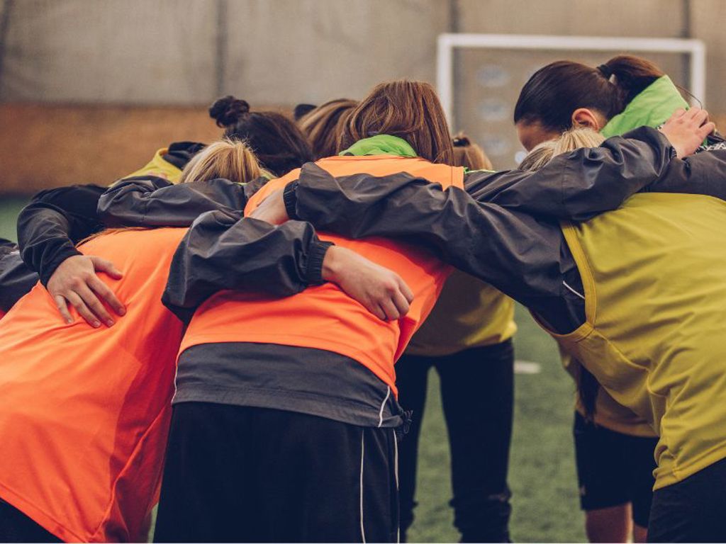 Tjejlag iklädda västar står i en ring med armarna om varandra på en konstgräsplan under tak