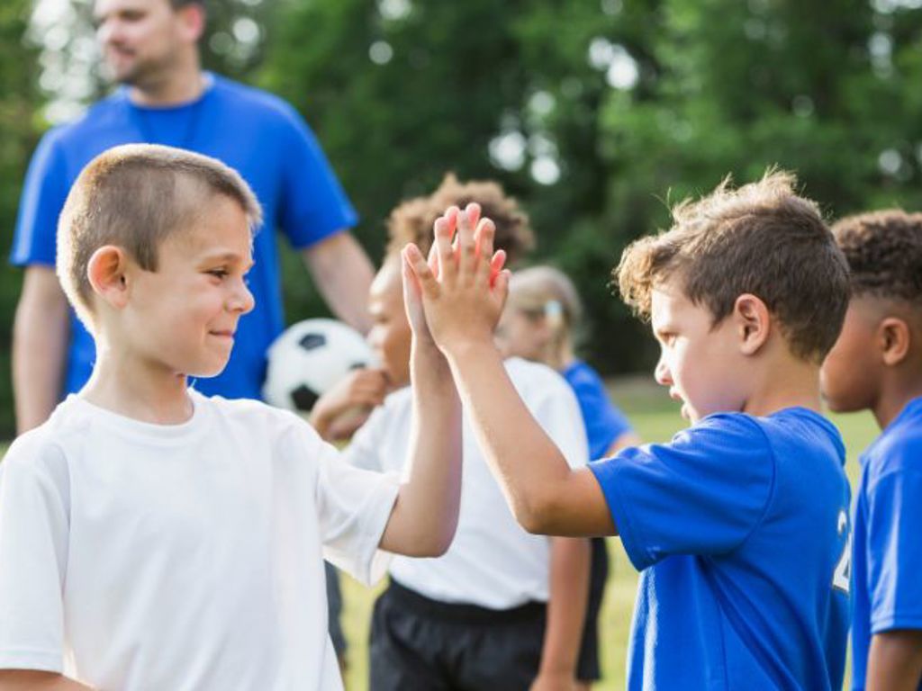 Pojkar i träningskläder ger varandra en high five, en tränare med en fotboll står i bakgrunden