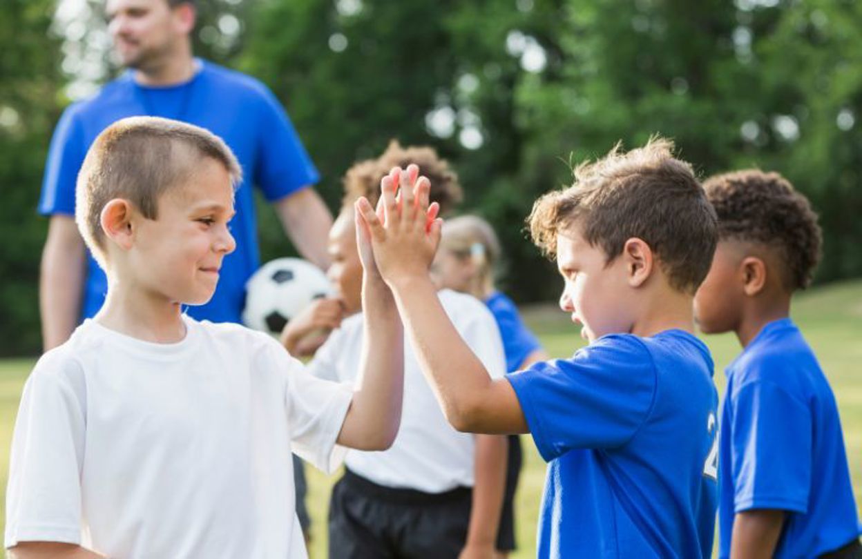 Pojkar i träningskläder ger varandra en high five, en tränare med en fotboll står i bakgrunden