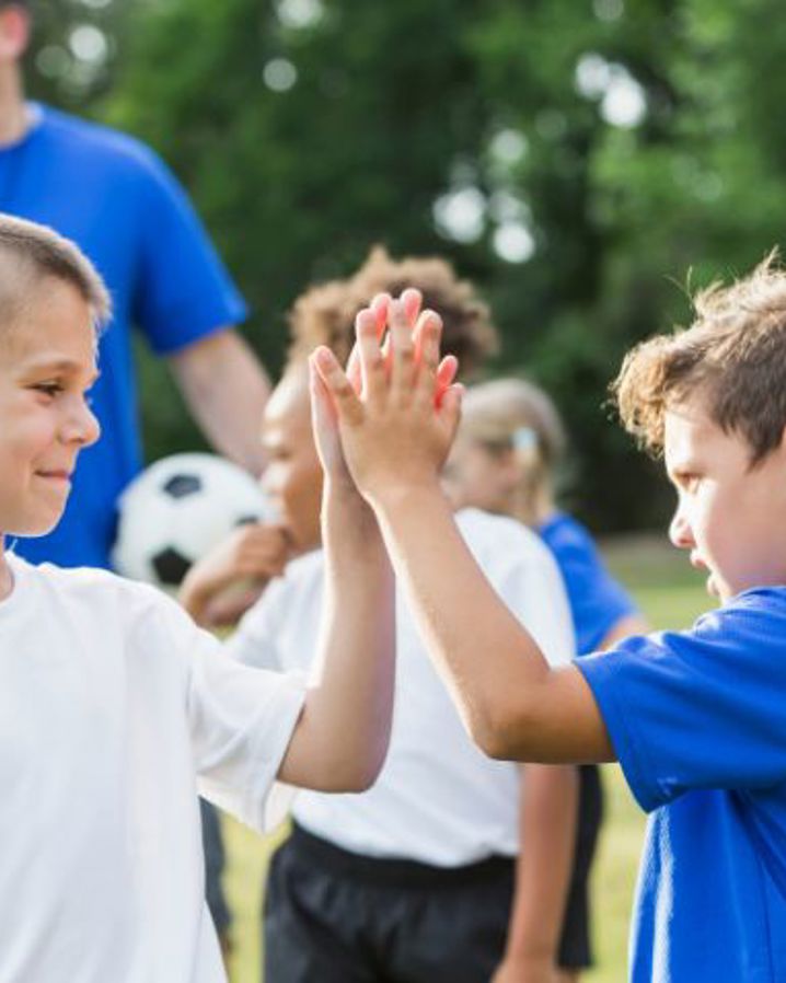 Pojkar i träningskläder ger varandra en high five, en tränare med en fotboll står i bakgrunden