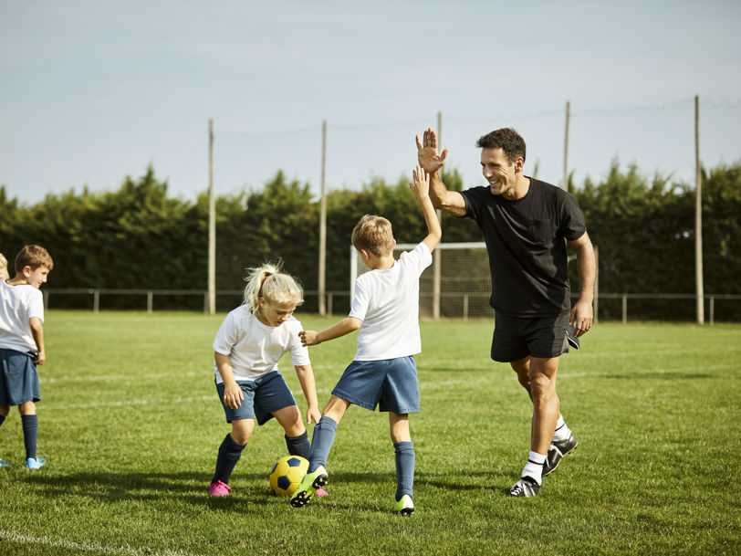 Barn på en gräsplan som tränar fotboll, ett utav barnen gör en high five med tränaren