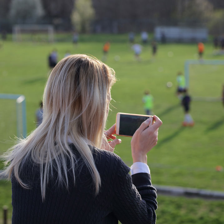 Kvinna sitter på en läktare framför en fotbollsplan och filmar träningen med mobiltelefonen.