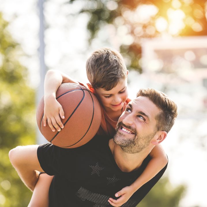 En man bär en pojke som håller en basketboll på ryggen i en grönskande utomhusmiljö