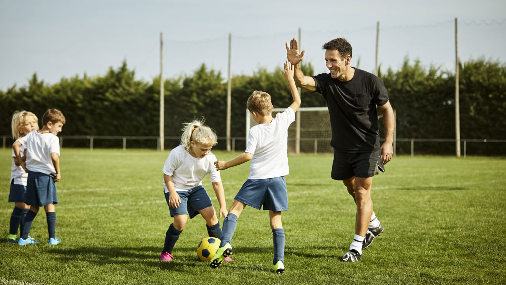 Barn på en gräsplan som tränar fotboll, ett utav barnen gör en high five med tränaren