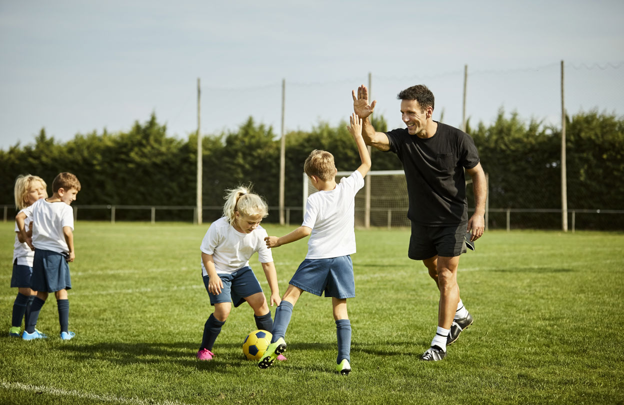 Barn på en gräsplan som tränar fotboll, ett utav barnen gör en high five med tränaren
