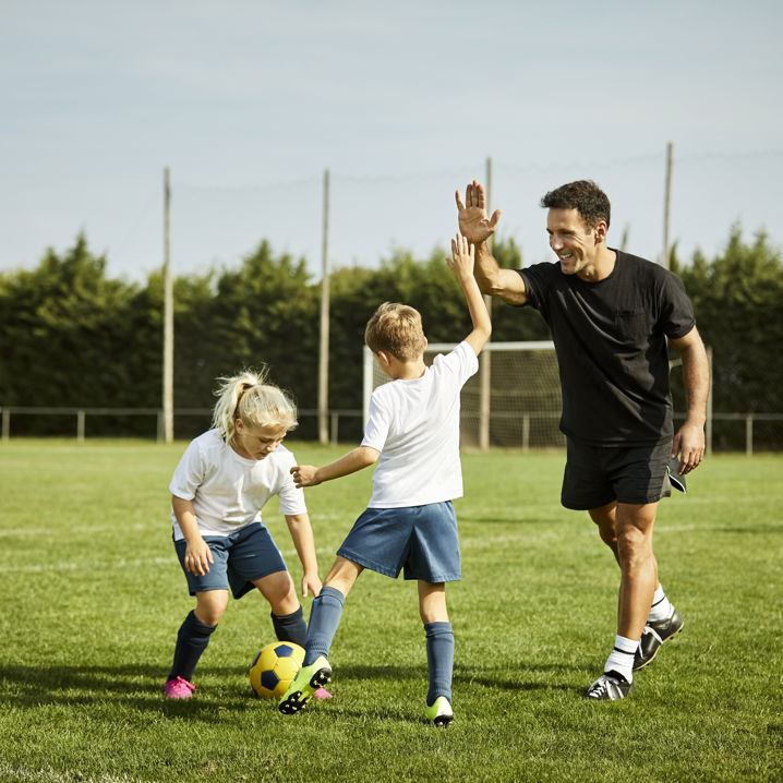 Barn på en gräsplan som tränar fotboll, ett utav barnen gör en high five med tränaren