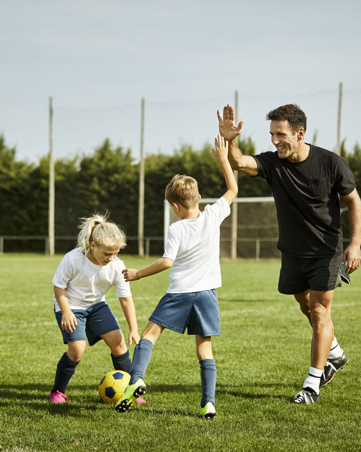 Barn på en gräsplan som tränar fotboll, ett utav barnen gör en high five med tränaren