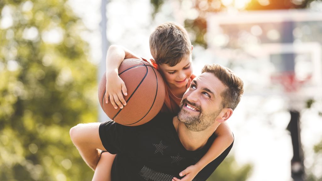 En man bär en pojke som håller en basketboll på ryggen i en grönskande utomhusmiljö