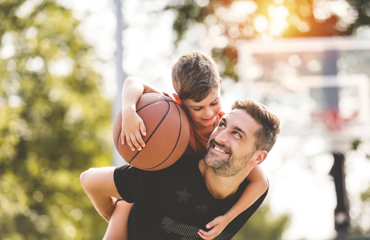 En man bär en pojke som håller en basketboll på ryggen i en grönskande utomhusmiljö