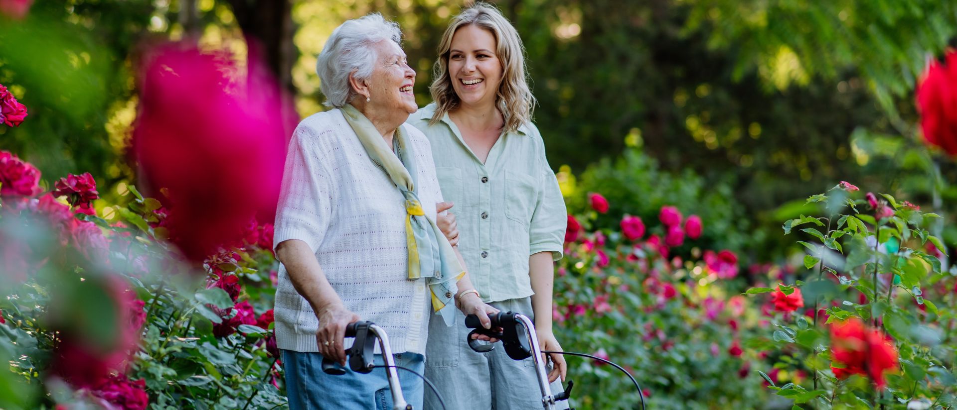 En yngre kvinna och en äldre kvinna med rullator promenerar på en trädgård full av blommor