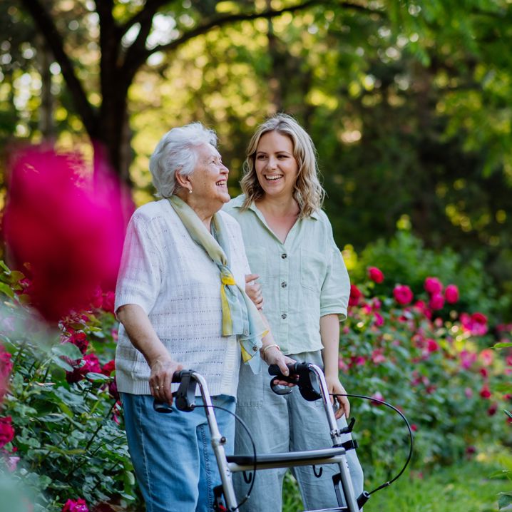 En yngre kvinna och en äldre kvinna med rullator promenerar på en trädgård full av blommor