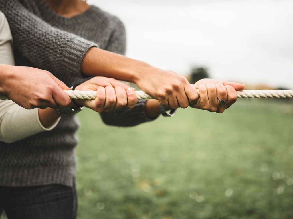 Två par händer som drar i ett rep som i en dragkamp i en grön utomhusmiljö