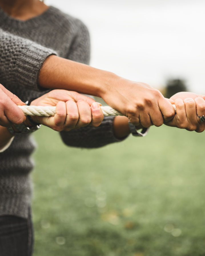 Två par händer som drar i ett rep som i en dragkamp i en grön utomhusmiljö
