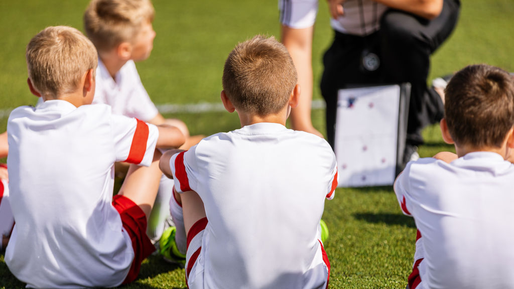 Fyra unga pojkar i fotbollskläder sitter med ryggarna mot kameran och tittar framåt mot en tränare med taktiktavla