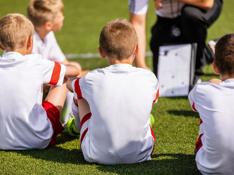 Fyra unga pojkar i fotbollskläder sitter med ryggarna mot kameran och tittar framåt mot en tränare med taktiktavla