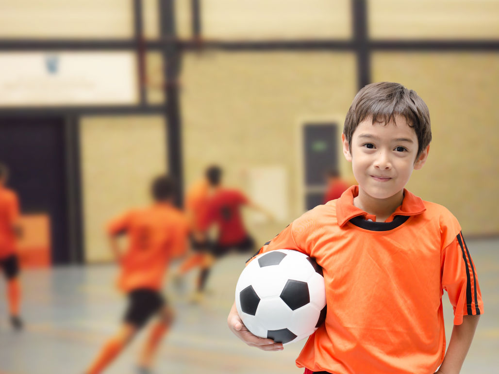 En pojke klädd i orange matchkläder och en fotboll under armen står i en idrottshall, med flera lagkompisar i bakgrunden