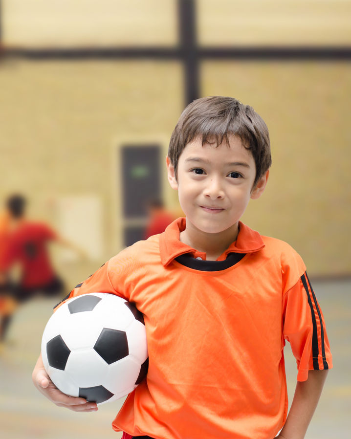 En pojke klädd i orange matchkläder och en fotboll under armen står i en idrottshall, med flera lagkompisar i bakgrunden