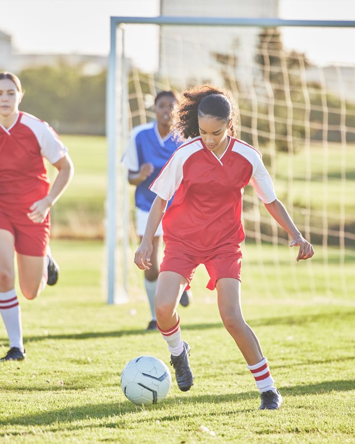 En tjej driver en fotboll från en gräsplan med tre andra spelare och ett fotbollsmål i bakgrunden