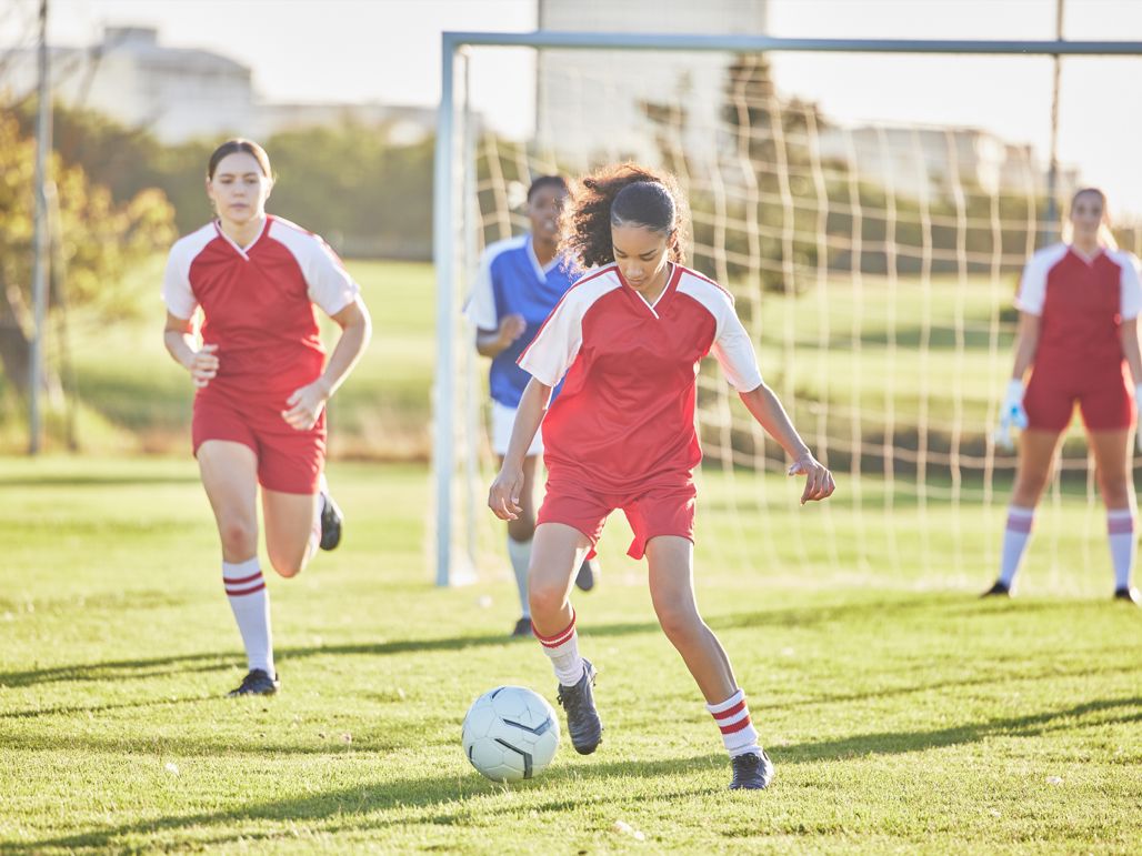En tjej driver en fotboll från en gräsplan med tre andra spelare och ett fotbollsmål i bakgrunden