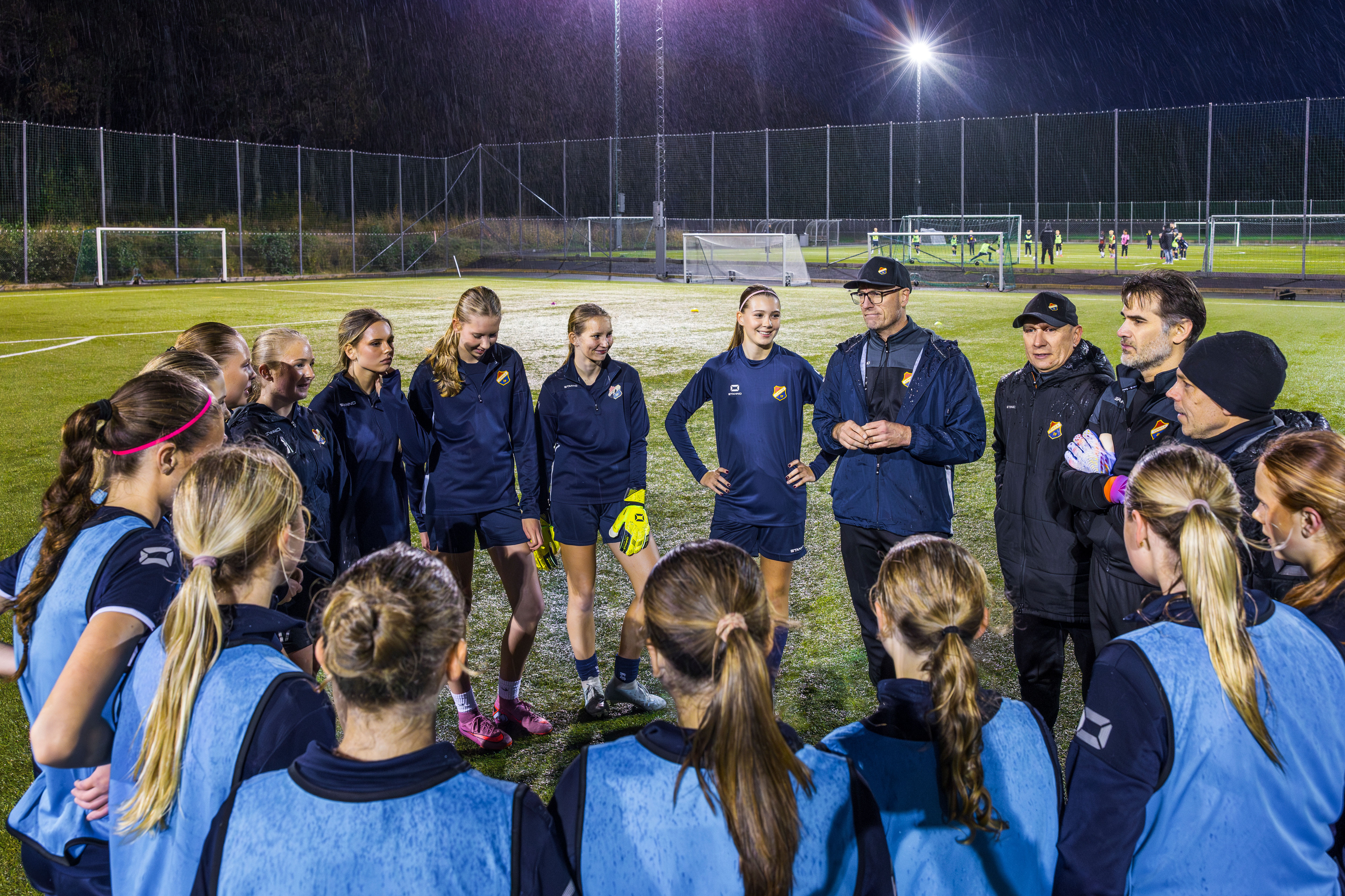 Kvällsbild på Lerkil F15 som samlas i ring på fotbollsplanen tillsammans med tränare inför match
