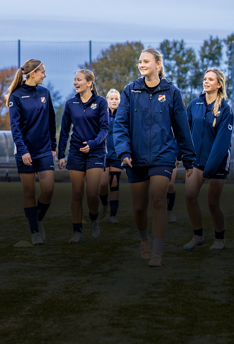 Fotbollstjejer med mörkblåa träningjackor umgås på en fotbollsplan i skymning