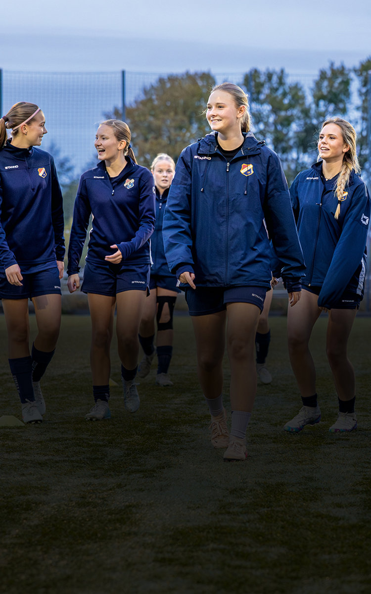 Fotbollstjejer med mörkblåa träningjackor umgås på en fotbollsplan i skymning