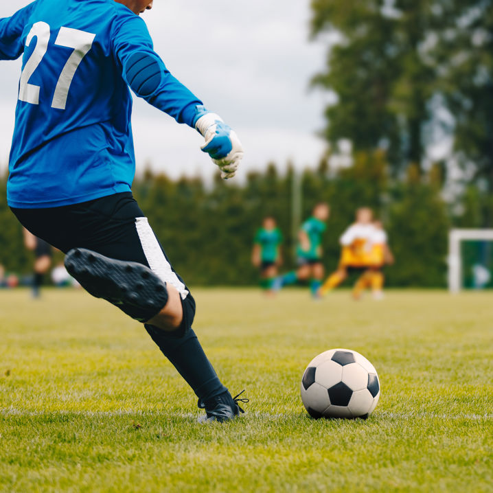 Fotbollsmålvakt på väg att skjuta ut bollen från liggande utspark, med med- och motspelare i bakgrunden
