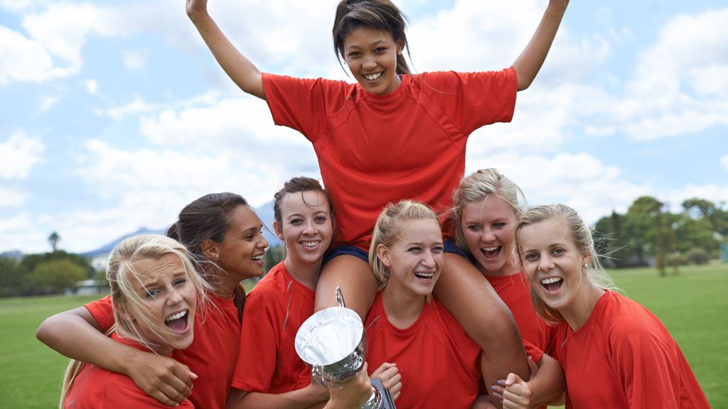 Tjejlag i fotboll med röda matchtröjor firar, med en fotboll och pokal i händerna. 