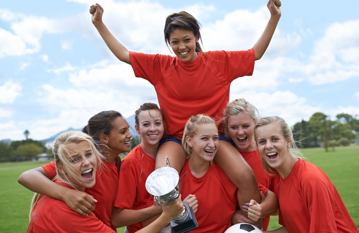 Tjejlag i fotboll med röda matchtröjor firar, med en fotboll och pokal i händerna. 