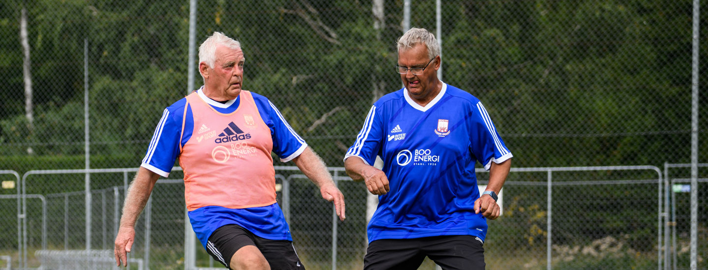 Ett par äldre herrar i blå tröjor, varav ena har en röd träningsväst, har en liten närkamp om fotbollen