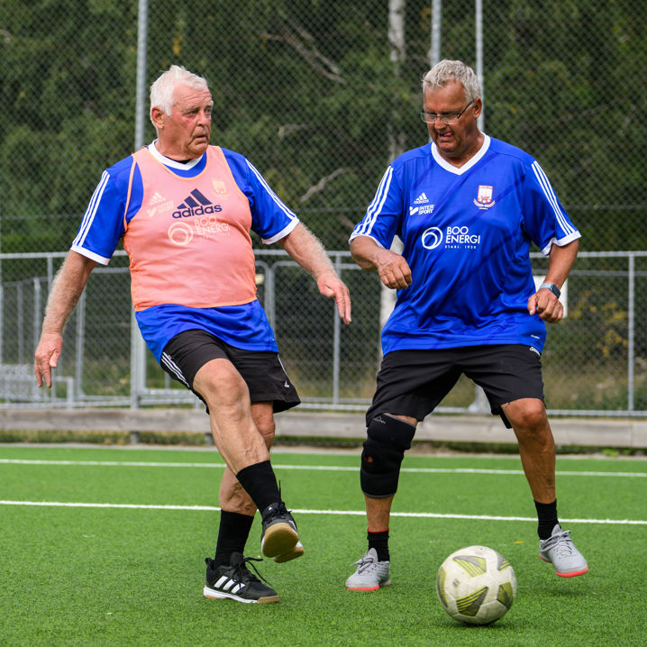 Ett par äldre herrar i blå tröjor, varav ena har en röd träningsväst, har en liten närkamp om fotbollen