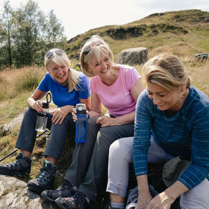 Tre skrattande kvinnor sitter på en kulle och fikar tillsammans