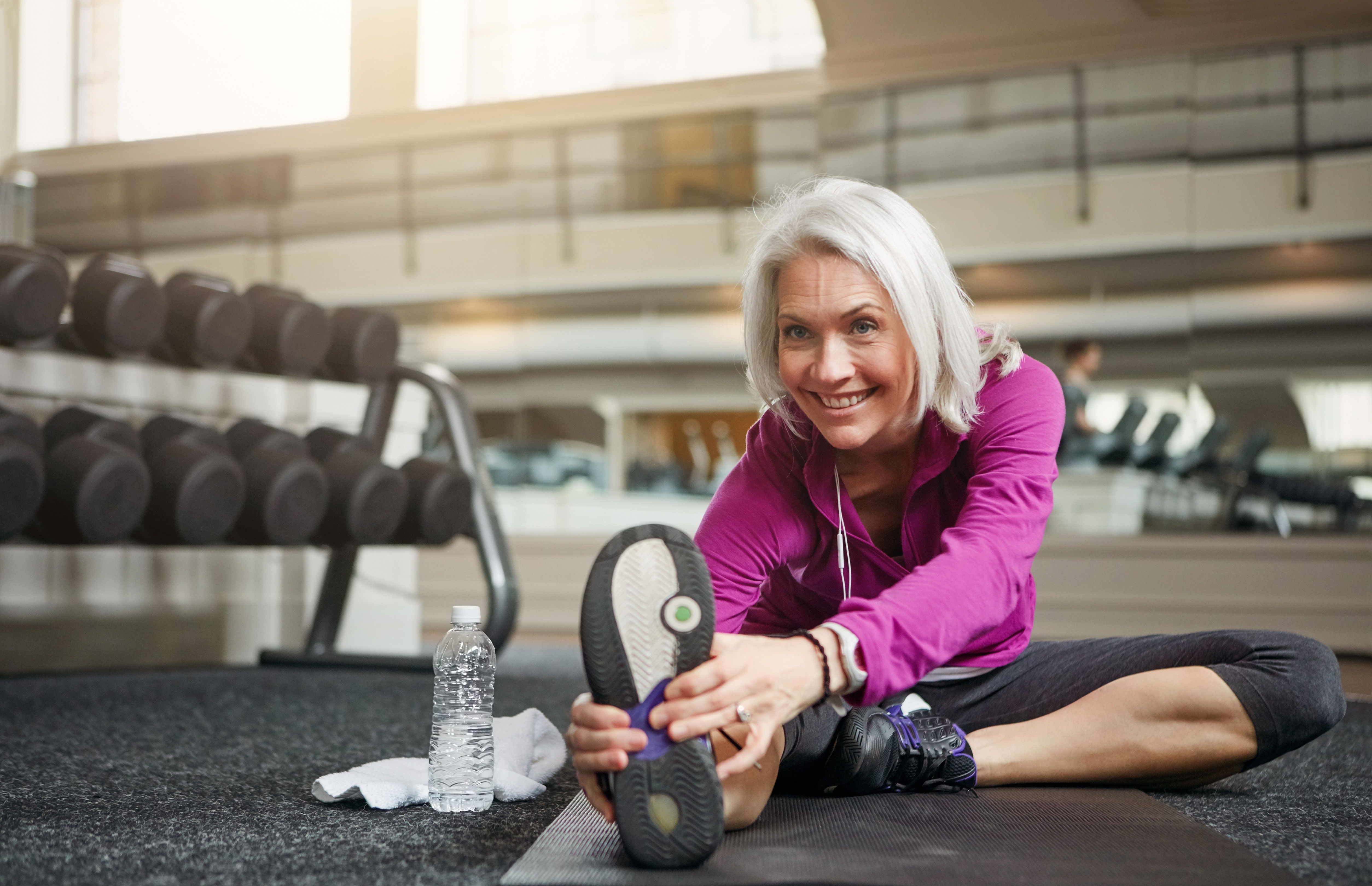 En äldre leende kvinna med grått hår och page i lila tröja som stretchar på gym