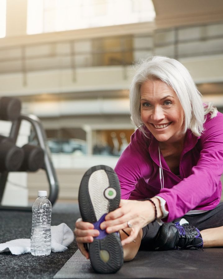 En äldre leende kvinna med grått hår och page i lila tröja som stretchar på gym