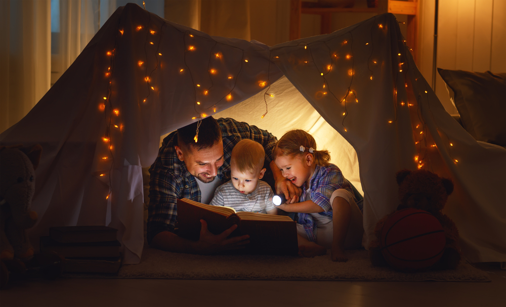 Pappa och barn som läser en bok med en ficklampa i ett tält hemma
