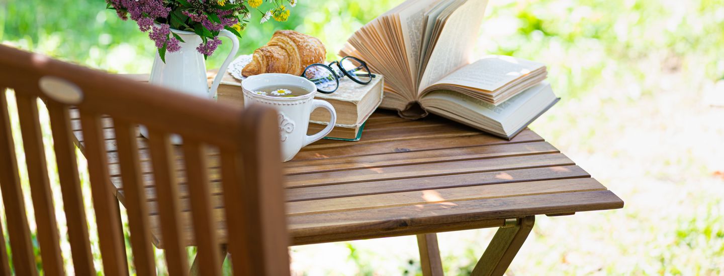 Ett bord i en grönskande trädgård, på vilket vi ser en bok, en kopp te, fika och en blomvas