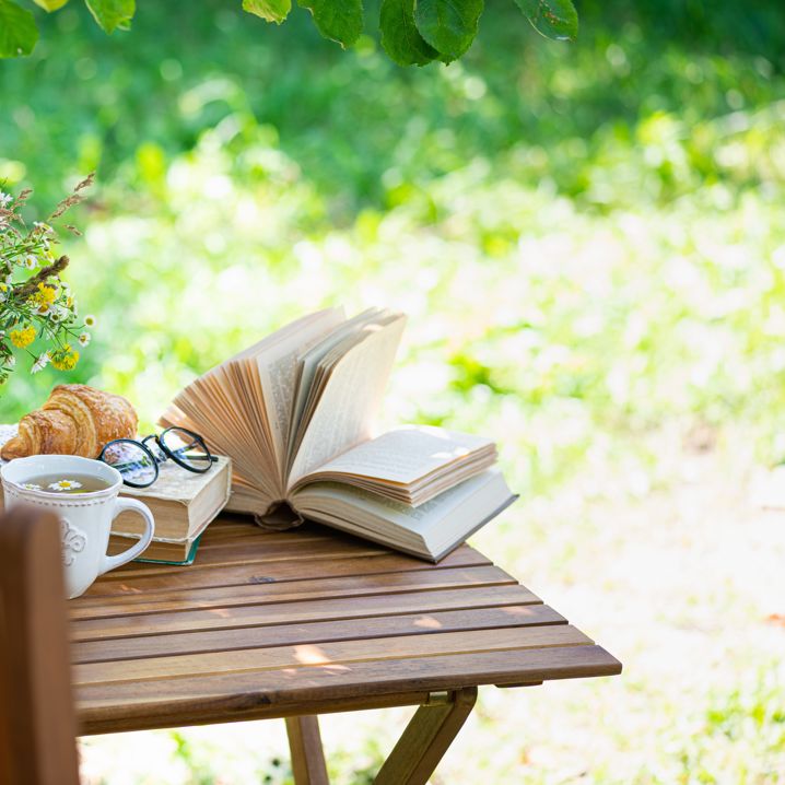 Ett bord i en grönskande trädgård, på vilket vi ser en bok, en kopp te, fika och en blomvas