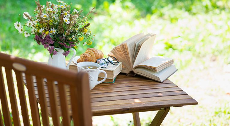 Ett bord i en grönskande trädgård, på vilket vi ser en bok, en kopp te, fika och en blomvas