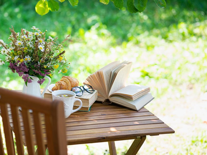 Ett bord i en grönskande trädgård, på vilket vi ser en bok, en kopp te, fika och en blomvas
